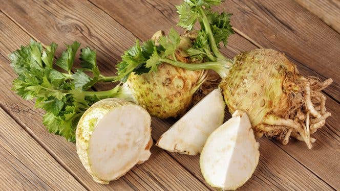 celeriac-for-website Two knobs of celeriac (celery root) on a wooden surface, with one knob cut into wedges showing the white flesh inside, and the other knob whole, with visible roots coming out of the bottom